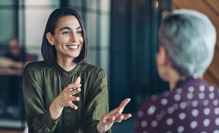 Two people engaged in conversation indoors. One, wearing a green shirt and gesturing with both hands, faces the camera with a blurred face. The other, seen from behind, has short gray hair and wears a polka-dotted shirt. Glass panels are visible in the background.